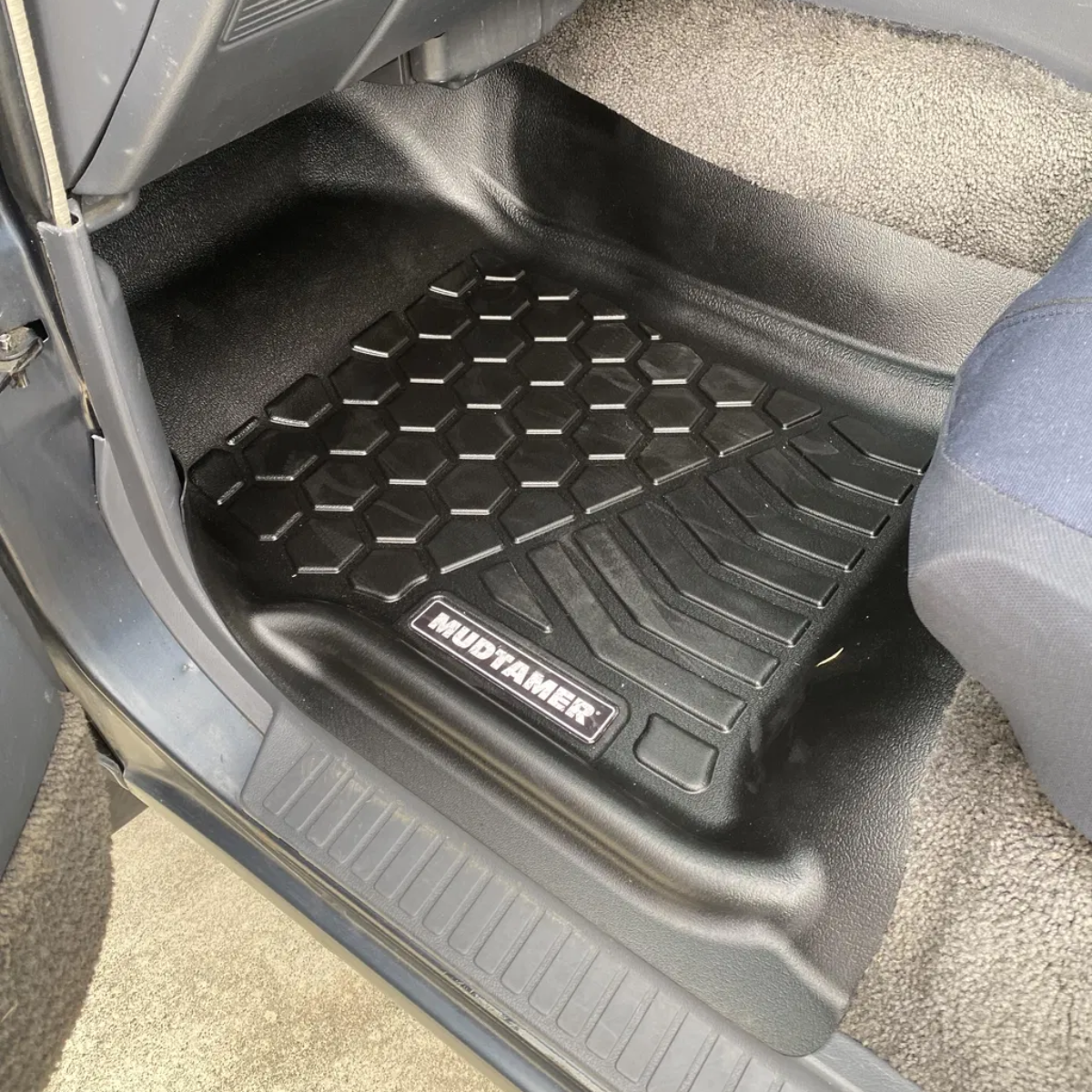 A black rubber car floor mat with a honeycomb pattern and a visible Mudtamer logo, shown installed inside a vehicle footwell next to a gray seat.